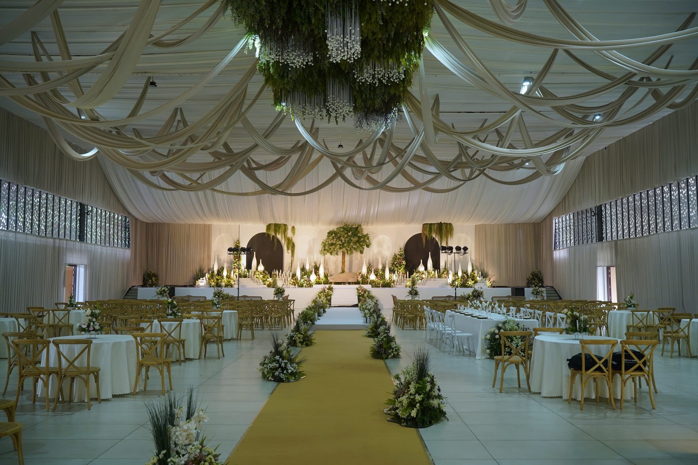 The ceremony aisle running beneath suspended greenery, chairs dressed in white